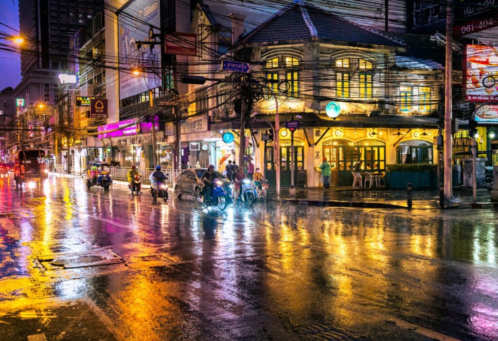 Explore the vibrant nightlife of Bangkok on a rainy evening, with bustling streets and neon lights reflecting in water.