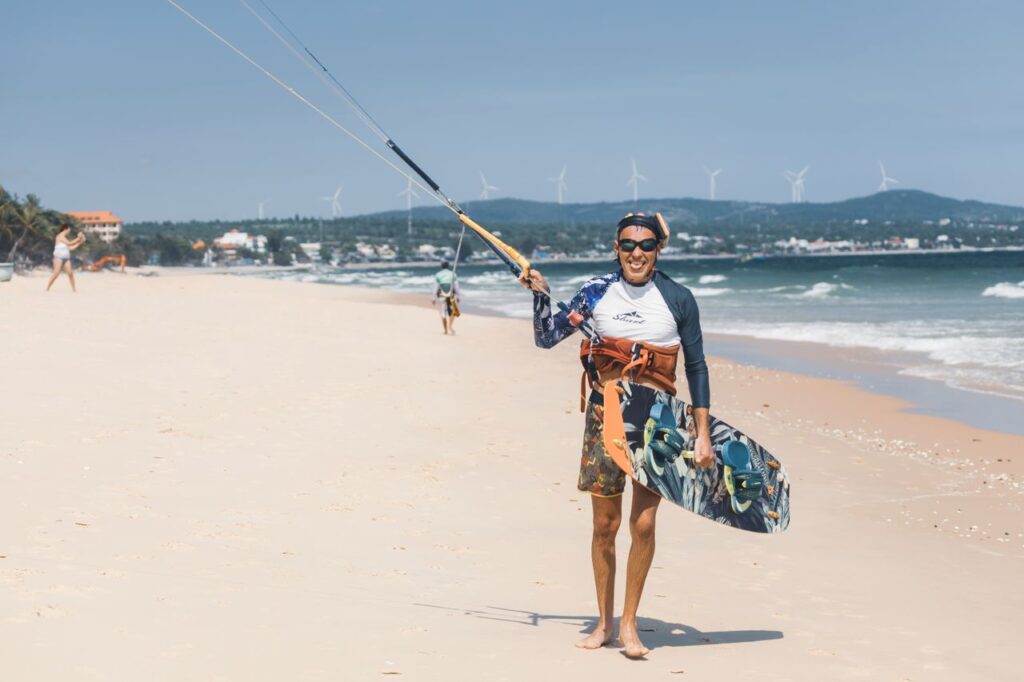 Smiling kitesurfer holding board on a sunny beach with wind turbines in the background.