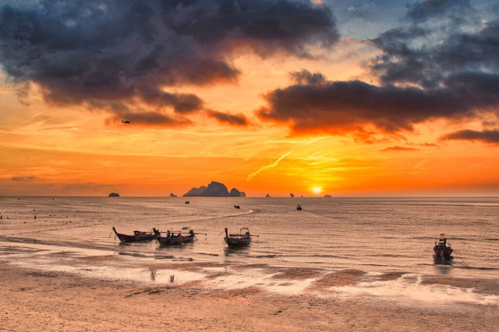 Golden sunset over Krabi beach with traditional boats and dramatic clouds.